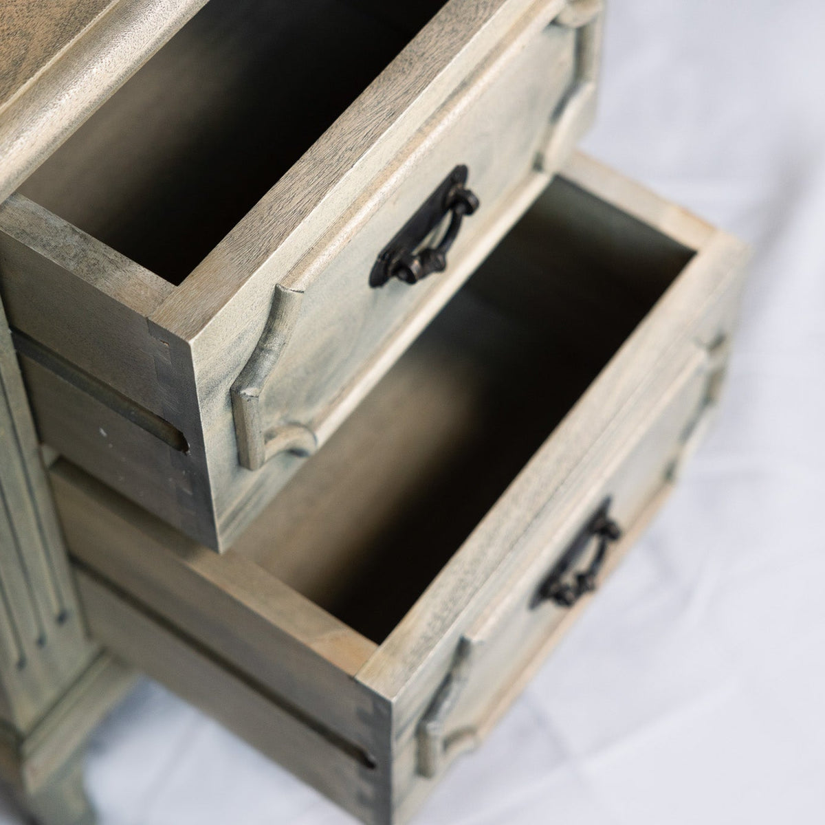 A close-up of a rustic two-drawer nightstand or side table with a distressed gray-green finish and dark metal drawer pulls, shown with both lower drawers open on a white fabric background.