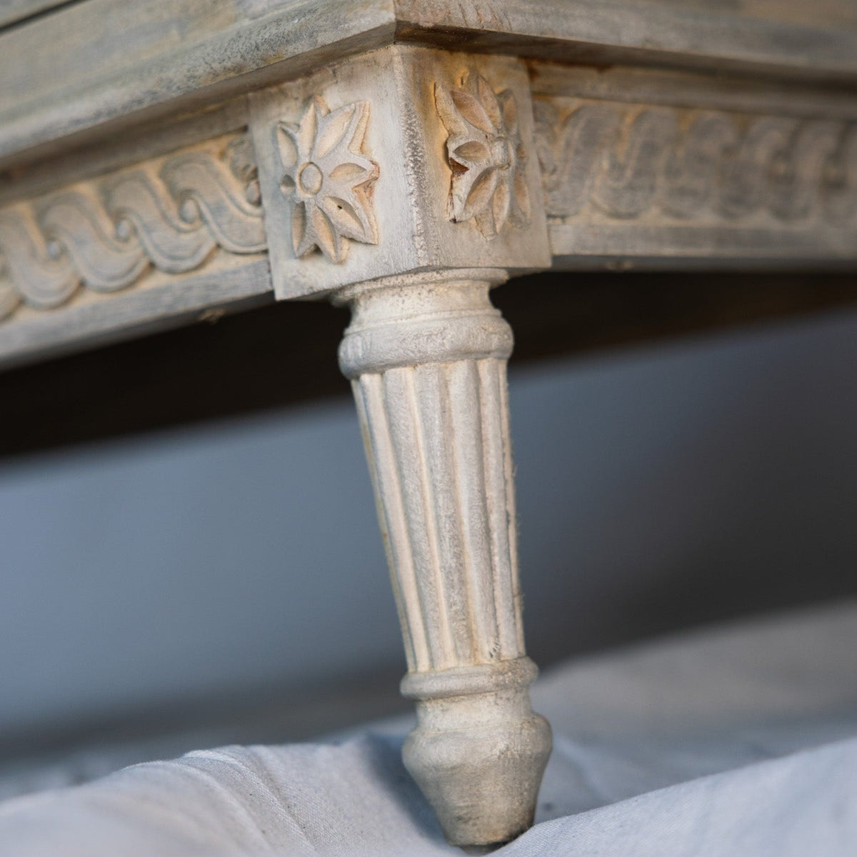 Close-up of an ornate distressed wood furniture leg with carved floral details and a fluted pedestal base, resting on white fabric.