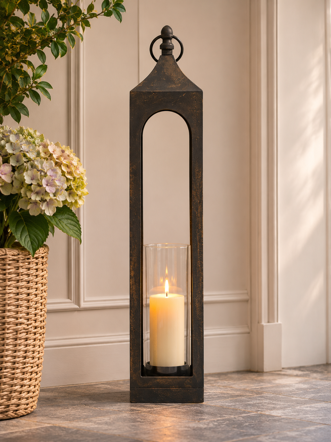 A tall black metal lantern-style candle holder with a clear glass cylinder and a lit white pillar candle inside, placed on a stone floor against a beige paneled wall with a wicker basket and flowers nearby.