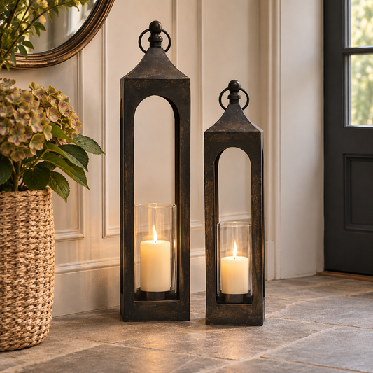 Two tall rectangular black metal lantern candles with arched cutouts and glass holders, placed on a stone floor in a home entryway beside a woven basket planter.