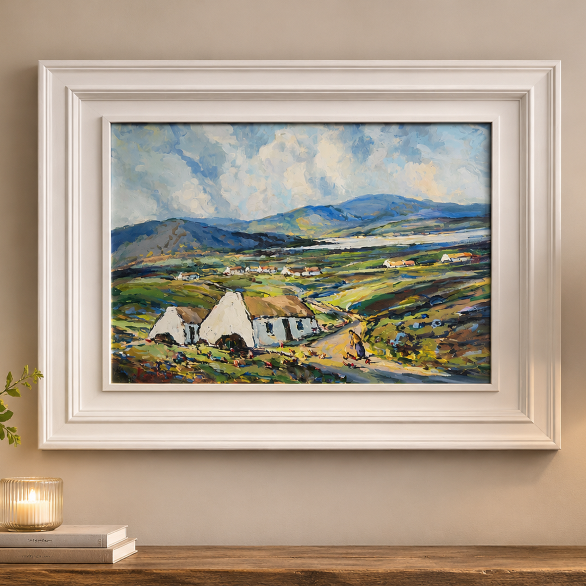 A framed landscape painting of white cottages in a green valley with mountains and a cloudy sky, displayed on a beige wall above a wooden console table with a lamp, candle, books, and flowers.