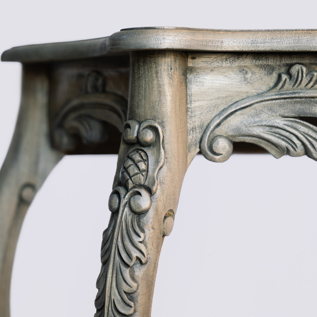 Close-up of an ornate carved wooden table leg with a distressed silver and gold finish, showing intricate scrollwork details against a plain light gray background.