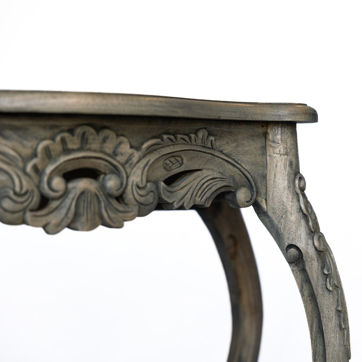 Close-up of an ornate carved wooden table corner with a distressed gray-brown finish, showing detailed cabriole-style legs and a tabletop edge against a plain white background.