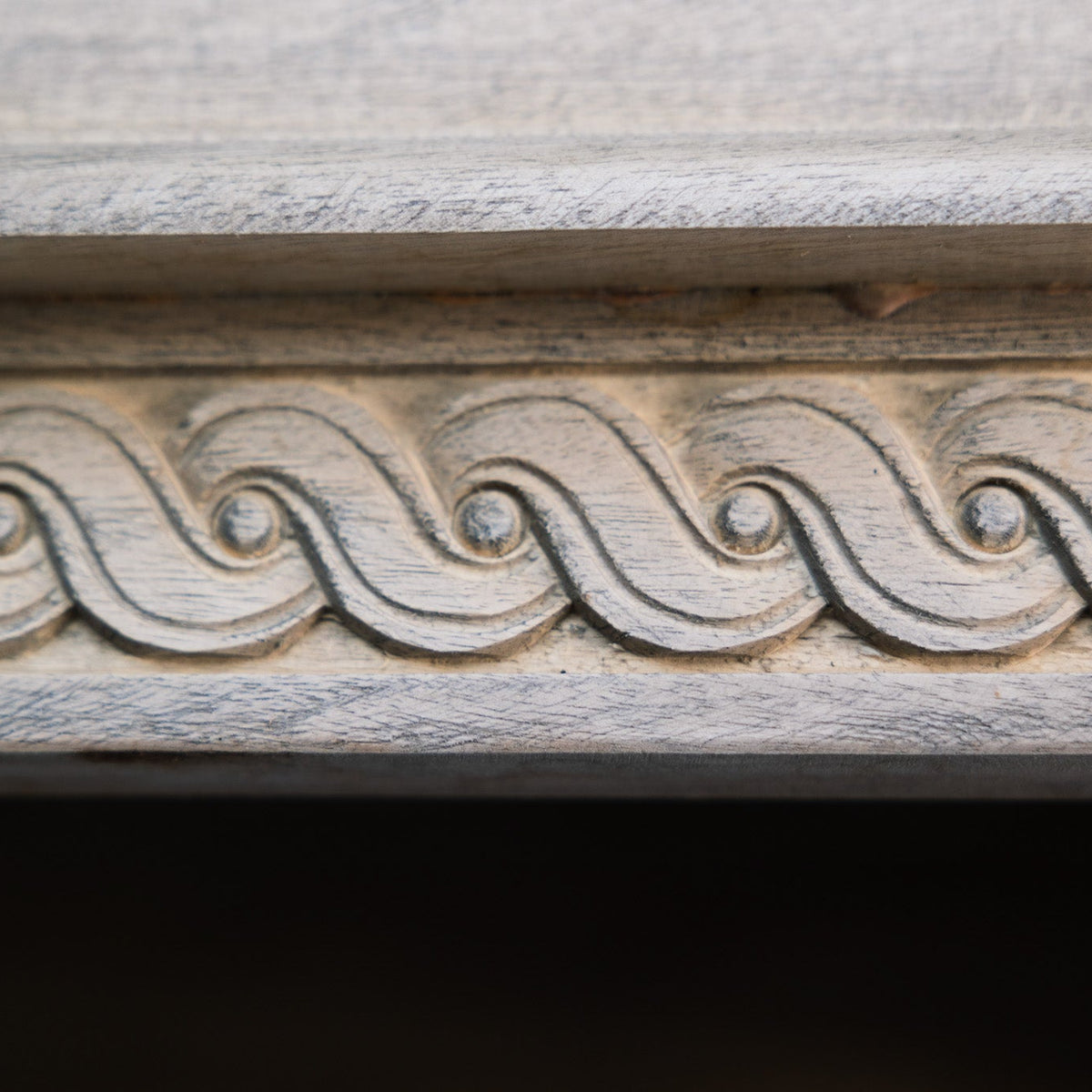 Close-up of a carved wooden trim with a repeating wave or scroll pattern, shown in weathered gray and beige tones against a dark background.