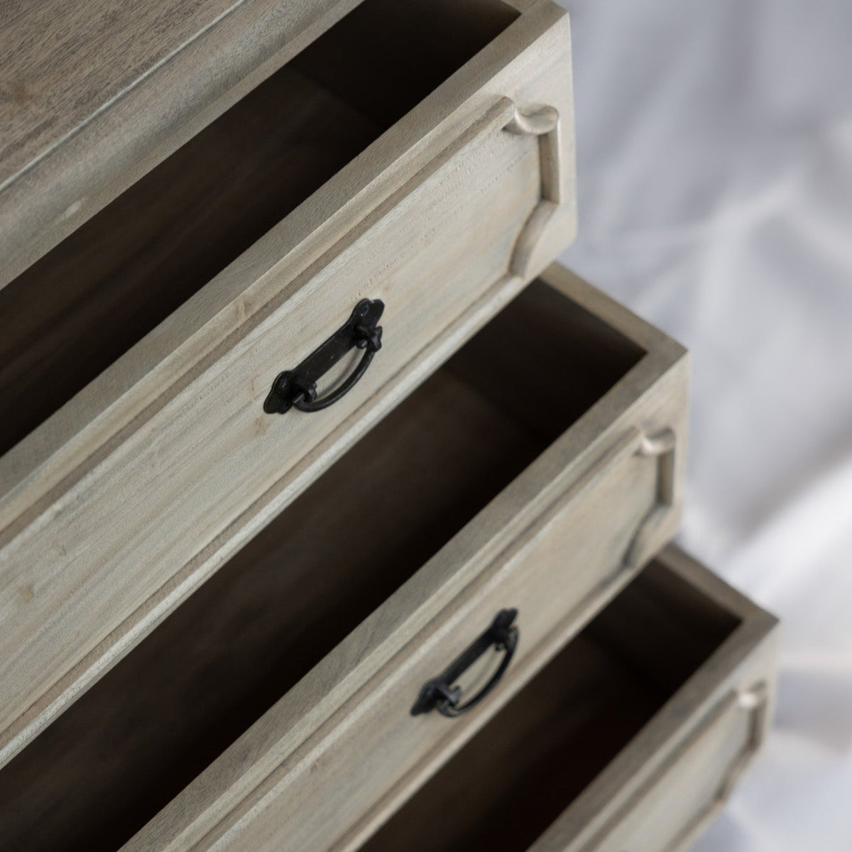 Close-up of a light natural wood dresser with three open drawers and black metal handles, set against a soft white fabric background.