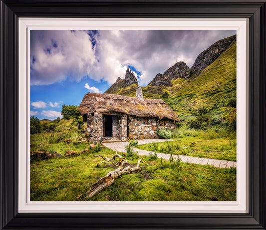 A framed landscape photograph of a rustic stone cottage with a thatched roof nestled in a grassy field, with dramatic mountains and a cloudy blue sky in the background. The print is matted with a white border and enclosed in a thick black frame.