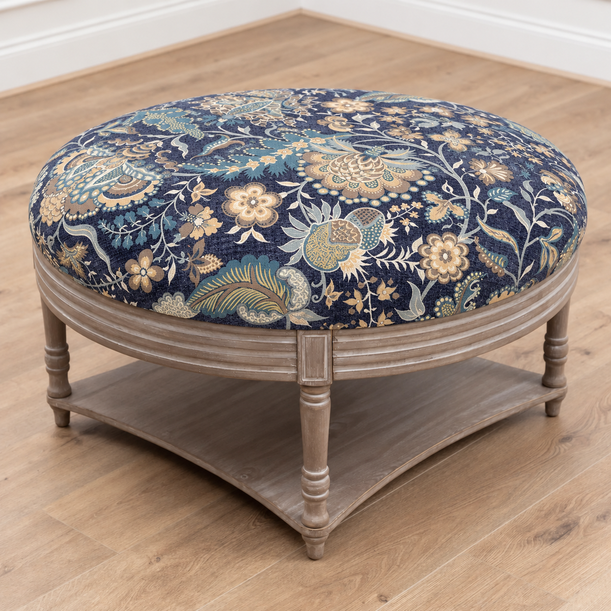 A round upholstered stool with a navy blue floral tapestry cushion and a distressed gray wooden frame, set on a light wood floor.