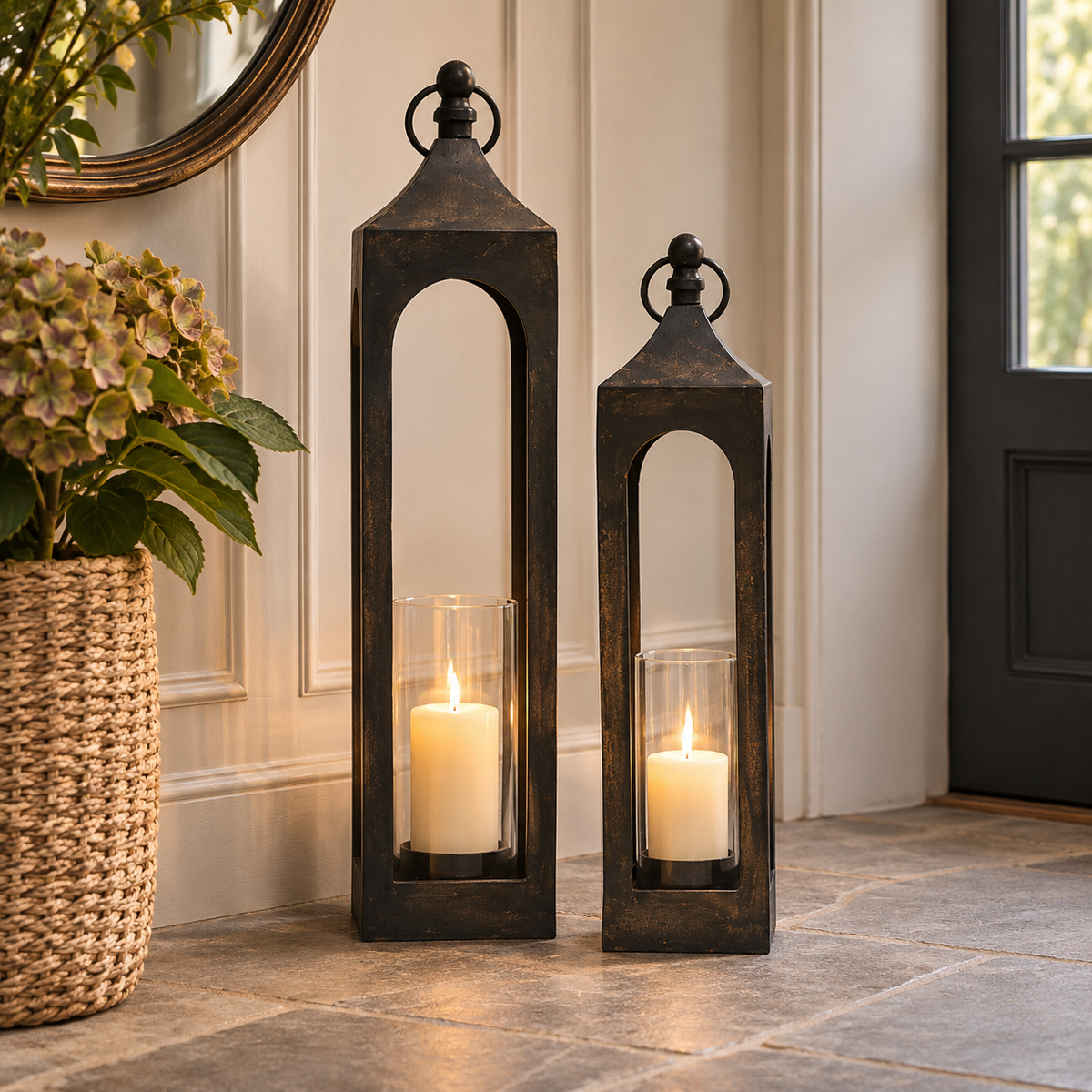 Two tall rectangular black metal lantern candles with arched cutouts and glass holders, placed on a stone floor in a home entryway beside a woven basket planter.