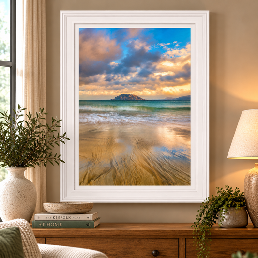A framed vertical art print of a beach and ocean sunset scene with dramatic clouds, displayed on a wooden console table in a warmly lit living room setting.