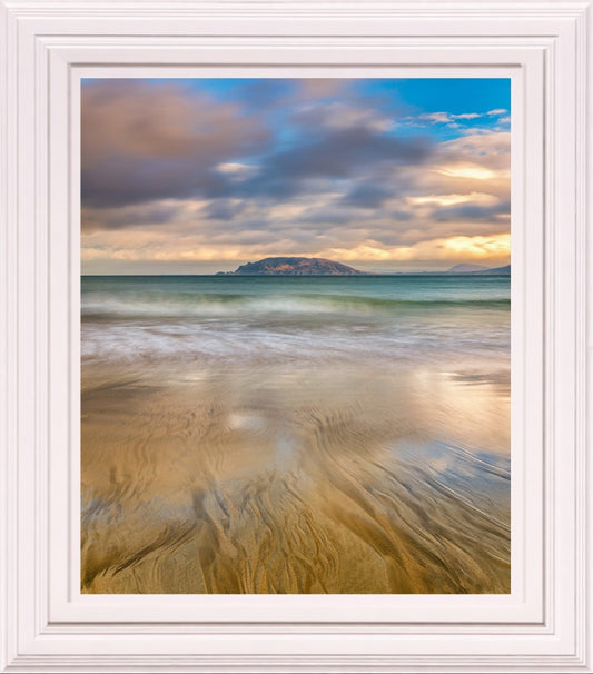 A framed photographic print of a beach seascape with wet sand in the foreground, calm water, a distant island, and a dramatic cloudy sky in blue, gray, and warm golden tones. The print is shown within a wide white frame against a white background.