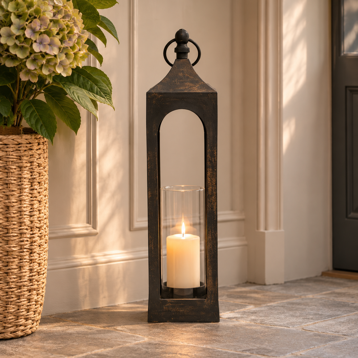 A tall black metal lantern-style candle holder with a glass cylinder insert and lit white pillar candle, placed on a stone floor against a cream wall with a woven planter and flowers nearby.