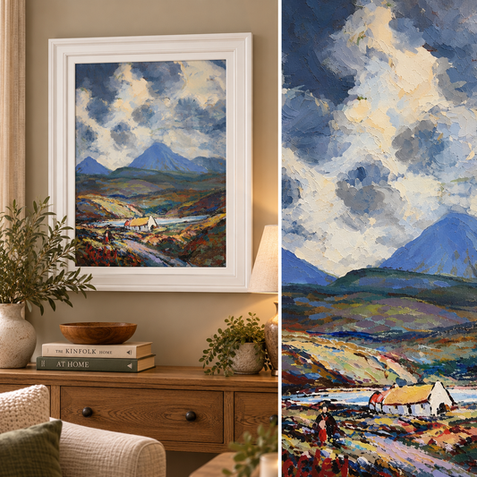 A framed landscape oil painting hangs on a beige wall above a wooden console table in a styled room. The painting depicts a dramatic mountain valley with blue mountains, a cloudy sky, and a small cottage.