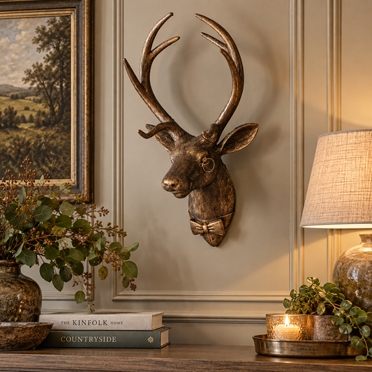 Bronze-toned metal stag head wall mount with antlers and a bow tie detail, displayed on a beige paneled wall above a wooden console table with books, a lamp, and plants.